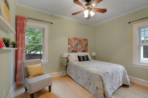 Bright main-floor bedroom with hardwood floors, a ceiling fan, and windows overlooking greenery.