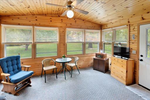 Beautiful wood-paneled porch with a matching wood ceiling, ceiling fan, and windows offering peaceful views of the mature trees and landscaped yard.