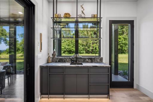 A stunning custom serving bar is highlighted by suspended iron-and-glass shelving, creating an open, airy focal point that beautifully showcases the surrounding landscape.