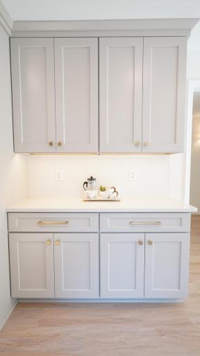 The light gray, factory-painted wood cabinetry and brushed gold hardware, staged here as a coffee bar, is fully custom with both ample storage and soft close doors.