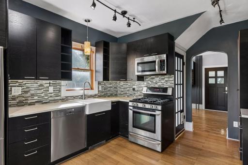 Bamboo flooring and track lighting complement the farmhouse sink, tile backsplash, and generous recycled resin countertop space. A window with a modern pendant light adds warmth and natural light.