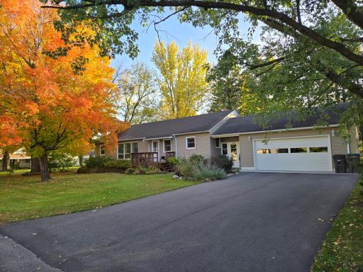 Inviting home surrounded by mature trees showcasing gorgeous fall colors, attractive landscaping, a welcoming front deck, and an attached two-car garage with a paved driveway.