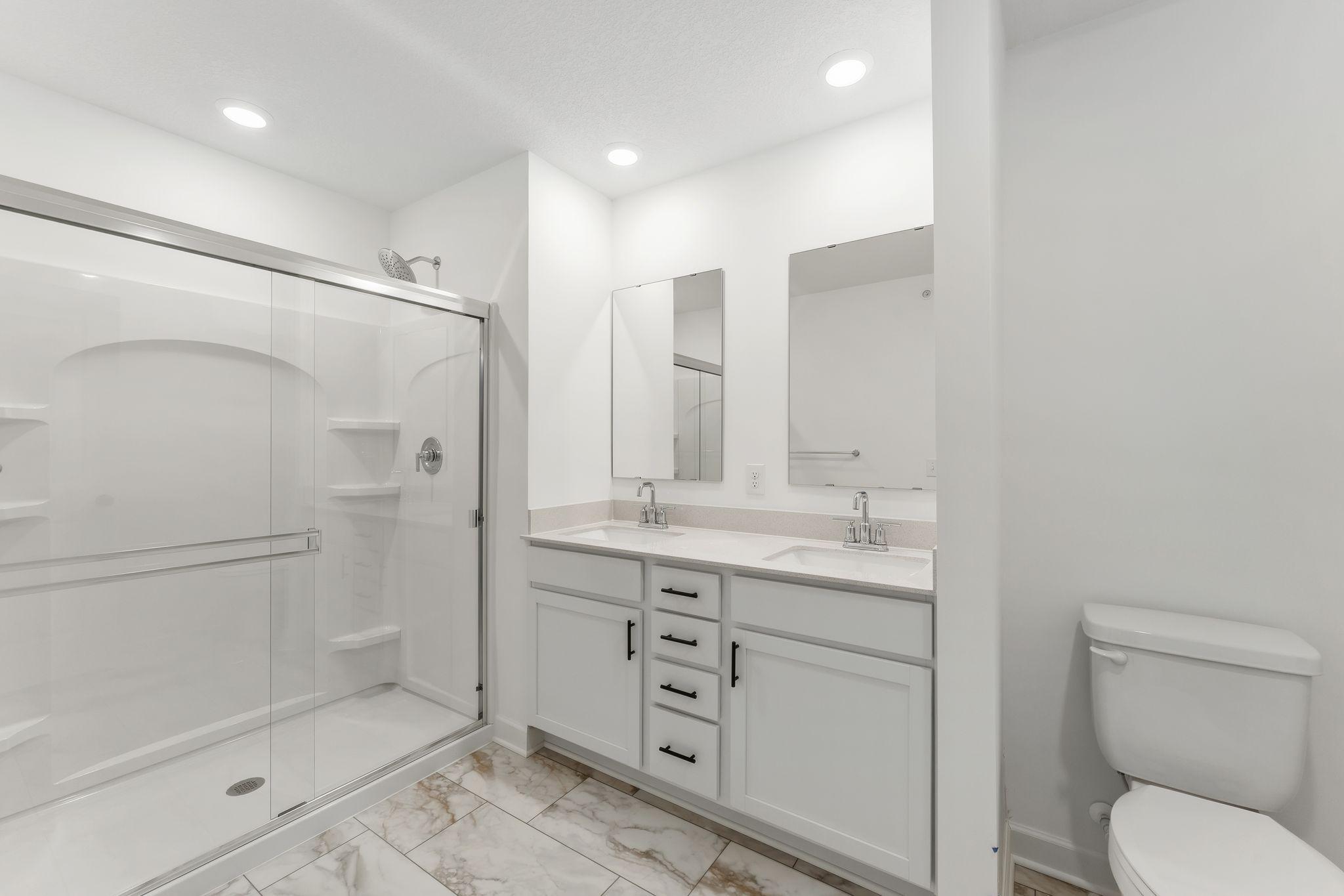 Featuring a glass-enclosed shower with built-in shelving and glass mirrors set atop dual quartz sinks and vanity-cabinetry, the en-suite bathroom helps streamline morning routines