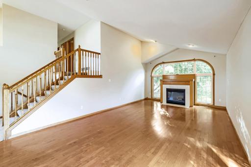 Light-filled great room with soaring vaulted ceilings and warm hardwood floors. Wood-mantel fireplace with tile surround anchors the space, framed by an arched window assembly overlooking green, treed views.