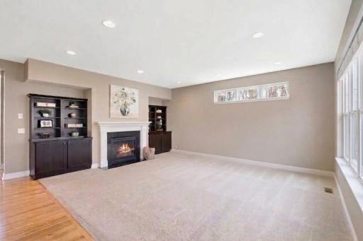 The focal point of the back wall is a classic fireplace with a white mantel, a dark firebox showing a warm flame, and framed artwork hanging above it. It is flanked by built-in dark wood cabinetry and shelving units.