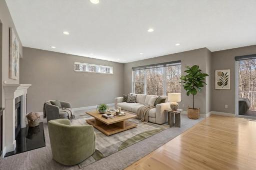 Spacious living room showcases abundant natural light, a fireplace, expansive windows with wooded views, and an open layout that flows seamlessly into the adjoining hardwood-floored space.