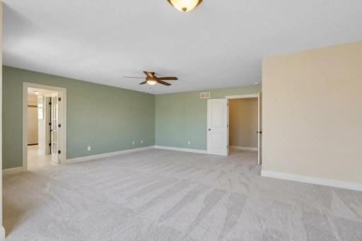 A second view highlights the impressive size and open layout of the primary suite, with a soft accent wall, ceiling fan, generous floor space, and direct access to the ensuite bath and adjoining areas