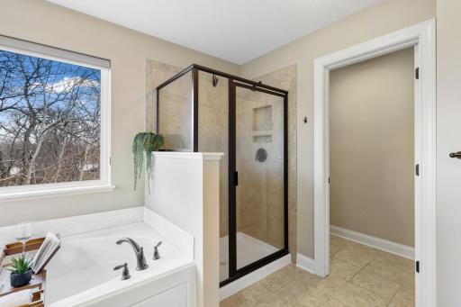 Spacious primary bath featuring a relaxing soaking tub and separate tiled walk-in shower with glass enclosure. A large window fills the space with natural light while offering peaceful wooded views, creating a bright and serene retreat