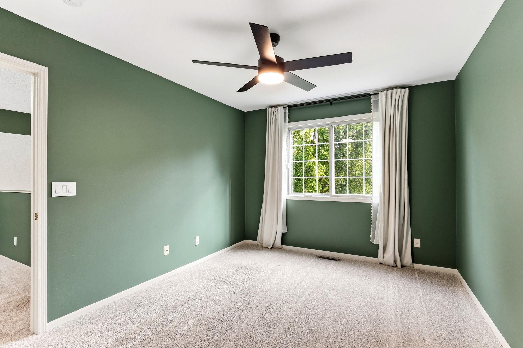 tep into the first bedroom, featuring a soothing professional green paint palette and a modern ceiling fan. A large window overlooks the tranquil neighborhood foliage, filling the room with natural light