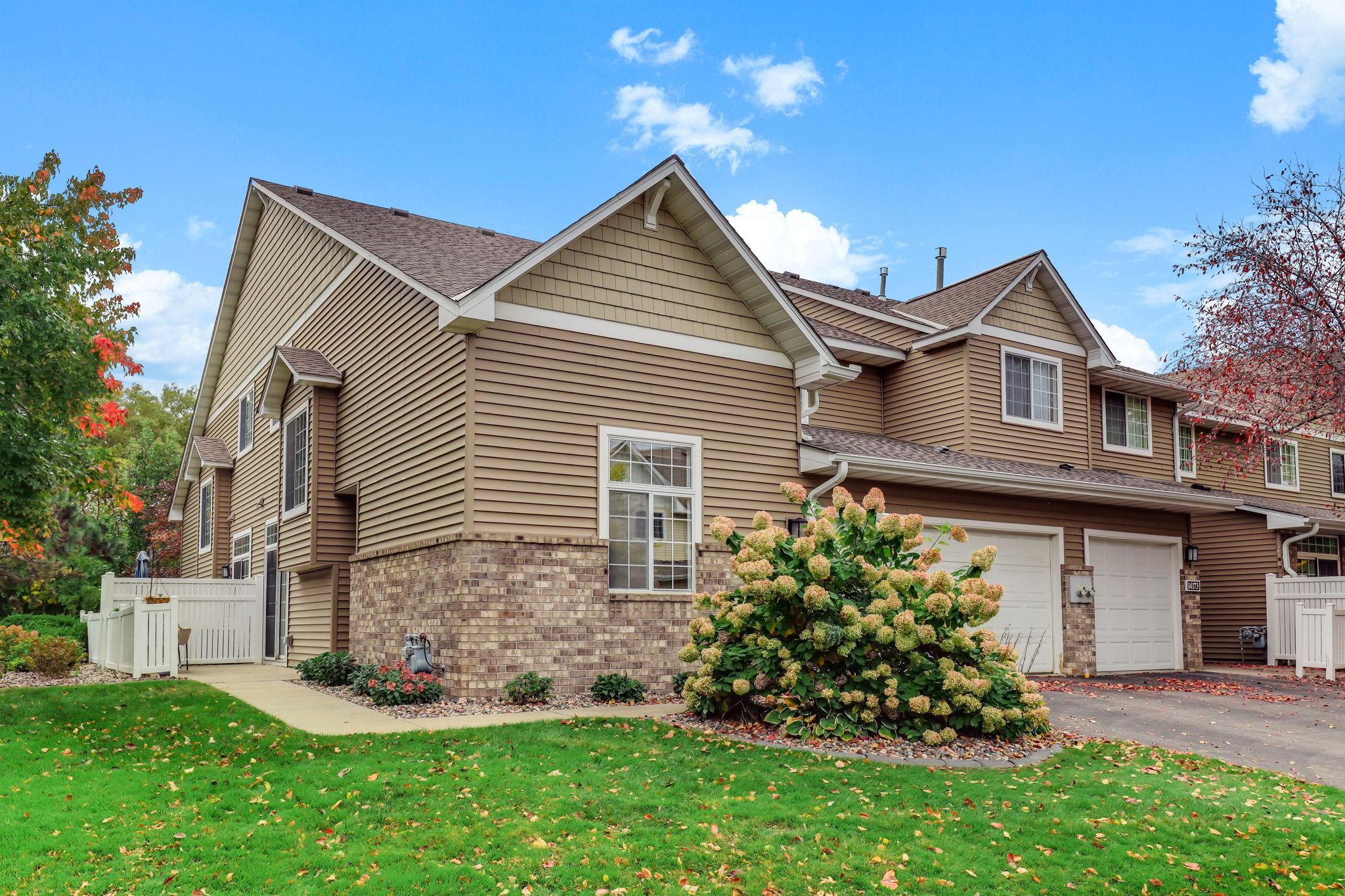 Welcome to this beautifully maintained end-unit townhome. The exterior features a classic mix of tan siding and brick accents, complemented by mature landscaping and a convenient attached garage.